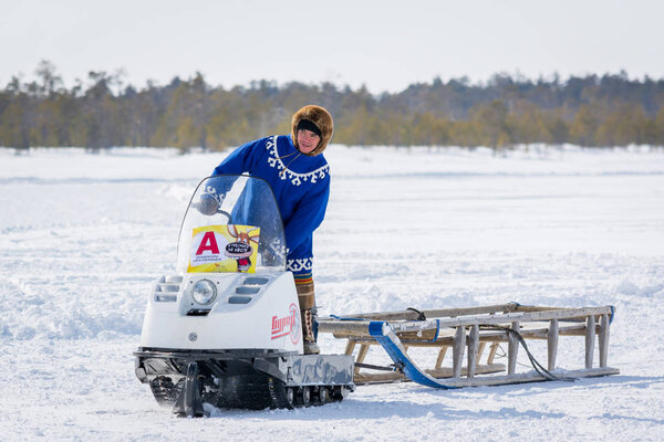 Russkinskaya, Russia - 24 March 2018: Taiga resident of siberia in national northern clothing, riding snowmobile with wooden sledges in the snow, against the background of winter forest. Holiday of th