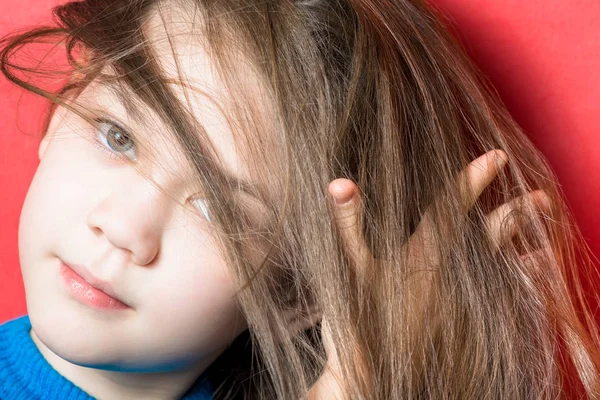 Cute girl in blue blouse tries to comb her hair with fingers on a red background