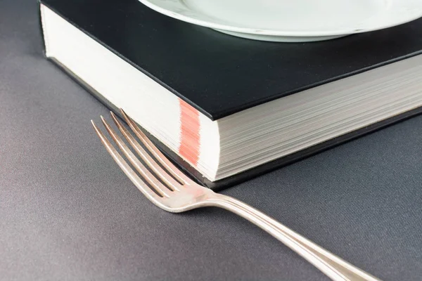 Λευκό πιάτο σε μαύρο βιβλίο δίπλα σε πιρούνι — Φωτογραφία Αρχείου