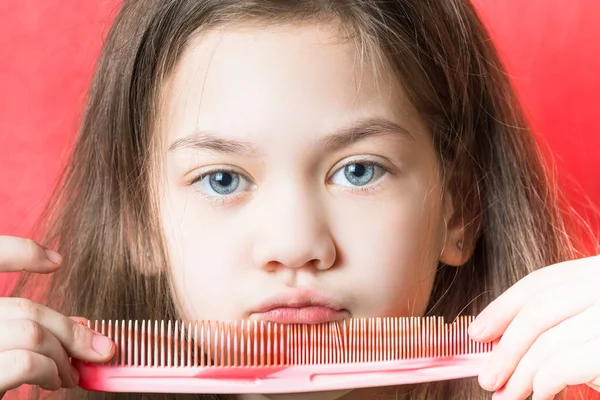 Cute girl holds a comb on her face against a red background — Stock Photo, Image