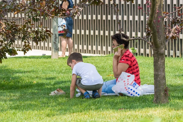 KRASNODAR, RUSSIE - 7 JUIN 2018 : Mère mature parlant sur son téléphone portable alors qu'elle est assise sur de l'herbe verte pendant que son enfant joue . — Photo