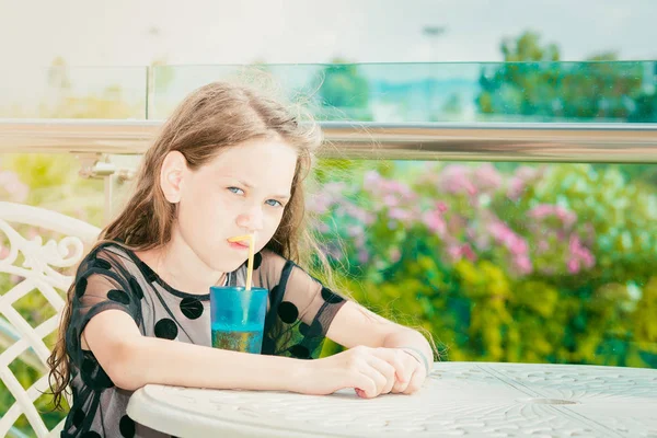 水玉模様の夏の黒いドレスを着た美少女がテーブルに座り、晴れた日には青いグラスでストローを通してジュースを飲んでいます。. — ストック写真
