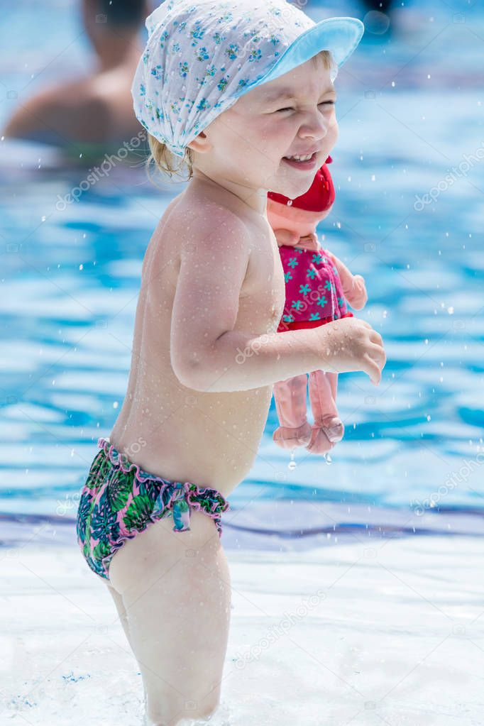 Beautiful child in shorts and cap with doll in his hand squints from sun in blue pool.