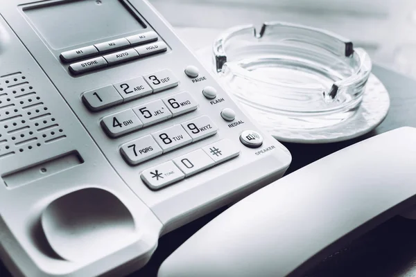 Λευκό τηλέφωνο με το τηλέφωνο κοντά στο γυάλινο τασάκι για κάπνισμα σε ξύλινο τραπέζι. — Φωτογραφία Αρχείου