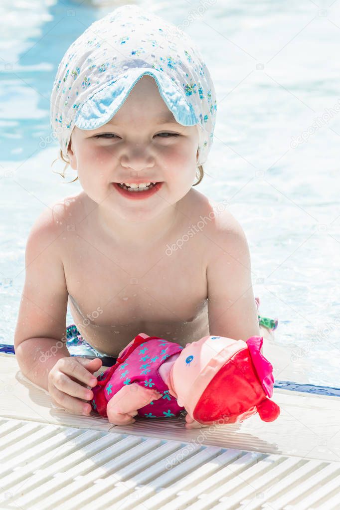 Small child with baby doll smiles in blue pool.