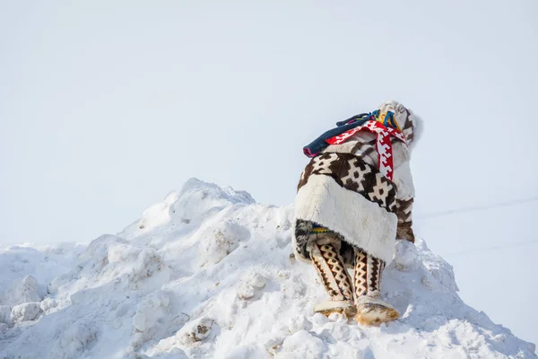 Russkinskaya, Rússia - 24 de março de 2018: Menino atarracado em roupas nacionais siberianas feitas de couro de pele de veado e pele, sobe na montanha nevada na praça central. Férias do pastor de renas . — Fotografia de Stock