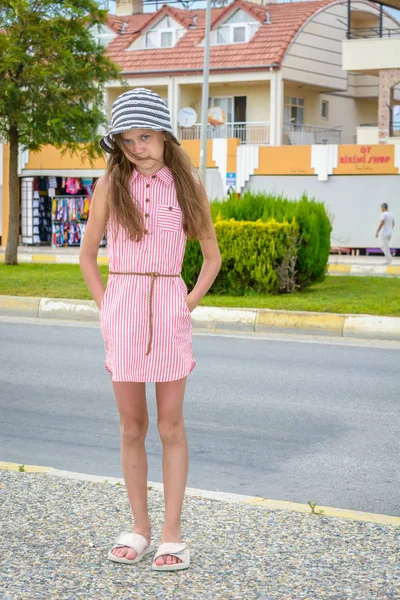 Lado, Turquía - Junio 2018: Una chica solitaria con sombrero y vestido de verano con las manos en los bolsillos está parada cerca de la carretera en medio de una gran casa . —  Fotos de Stock