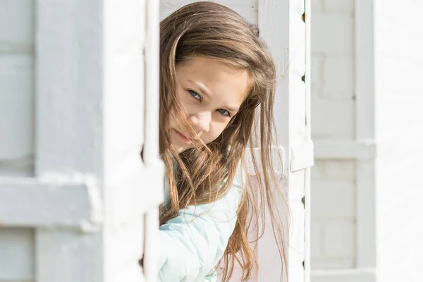 青いダウンジャケットで流れる髪を持つシャギーの女の子は、白いレンガの柱の後ろから外を見ます. — ストック写真