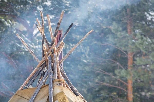 Den övre delen av vinter bostaden i norra folk med trästolpar insvept i renskinn och pipa från spisen mot skogens bakgrund — Stockfoto