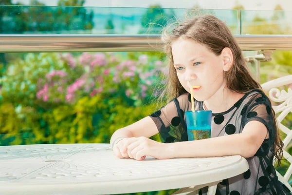 水玉模様の夏の黒いドレスを着た美少女がテーブルに座り 晴れた日には青いグラスでストローを通してジュースを飲んでいます — ストック写真