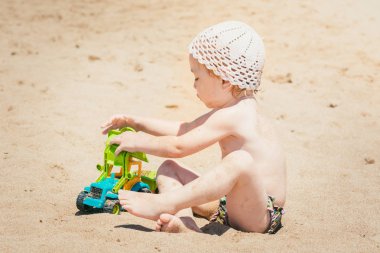 Kumlu plajda oyuncak traktör ile oynarken beyaz şapkalı güzel bebek