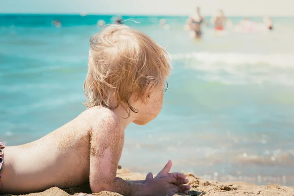 Kleines Kind liegt und sonnt sich auf heißem Sand unter Sonnenstrahlen und betrachtet Badende im Meer. — Stockfoto