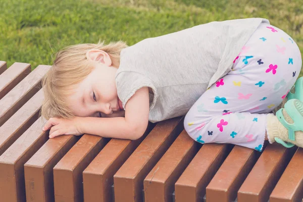 明るい服を着た満足した子供は、市内の公園の長いベンチで横たわって休んでいる. — ストック写真