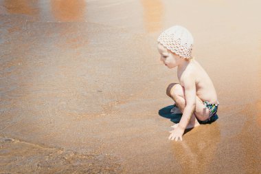 Yaz güneş şapka memnun çocuk kumlu plaj kıyısında oturur ve su ile oynar.
