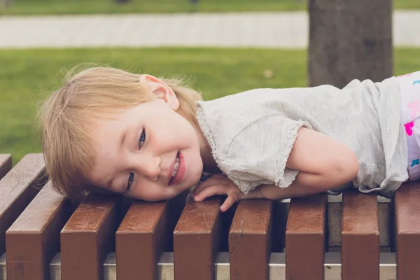 明るい服を着た満足した子供は、市内の公園の長いベンチで横たわって休んでいる. — ストック写真