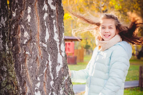Lächelndes Mädchen mit zerzausten Haaren im Wind in Schal und blauer Daunenjacke hält sich an einem Herbsttag an Birke fest. — Stockfoto