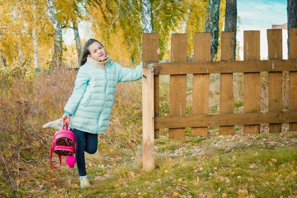 Grande fille en écharpe et veste en duvet bleu tient sac à dos de ville rouge par clôture en bois dans le village par une journée fraîche d'automne . — Photo