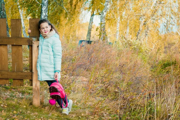 Grande fille en écharpe et veste en duvet bleu tient sac à dos de ville rouge par clôture en bois dans le village par une journée fraîche d'automne . — Photo