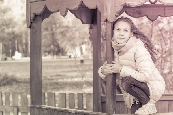 Nettes Mädchen in Daunenjacke sitzt an einem Herbsttag auf einem Pavillon. — Stockfoto