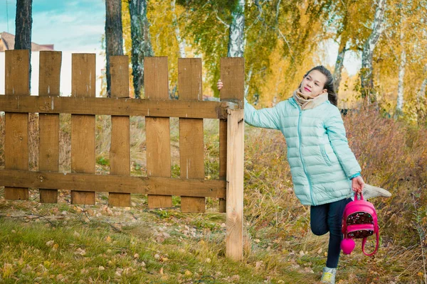 スカーフと青いダウンジャケットの背の高い女の子は、秋の涼しい日に村の木製のフェンスで赤い都市のバックパックを保持しています. — ストック写真