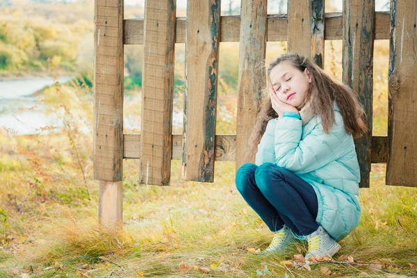 スカーフと青いダウンジャケットのかわいい女の子は、秋の涼しい日に村の木製のフェンスで閉じた彼女の目で眠ります. — ストック写真