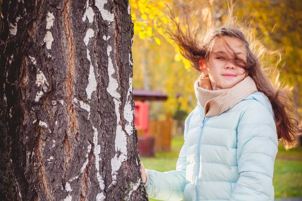 スカーフと青いダウンジャケットで風の中で散らばった髪を持つ笑顔の女の子は、秋の日にバーチにつかまっている. — ストック写真