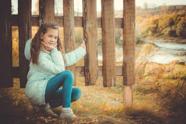 スカーフと青いダウンジャケットの美しい女の子は、秋の涼しい日に村で木製のフェンスを持ってしゃがんでいます. — ストック写真