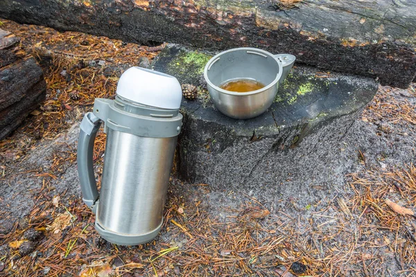 Thermos de camping en plein air avec tasse de thé se tient sur souche dans la forêt . — Photo