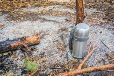 Sıcak çay veya kahve için çelik termos soyu tükenmiş şenlik ateşinin yanında ormanda yerde.