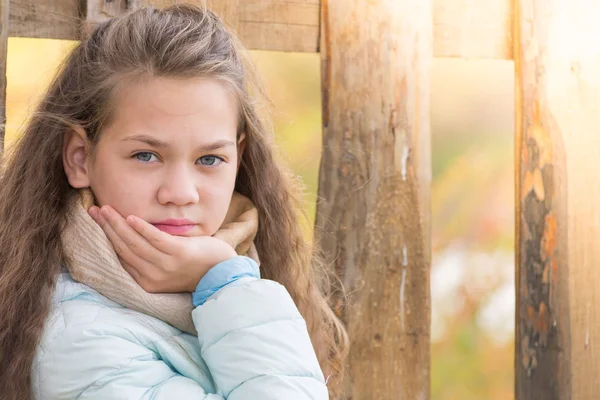 スカーフと青いダウンジャケットのかわいい女の子は、秋の涼しい日に村の木製のフェンスの背景に彼女の手を維持します. — ストック写真