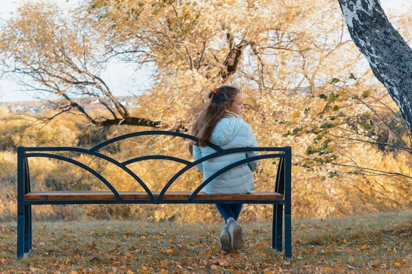 悲しい気分のライトダウンジャケットの美少女は、涼しい秋の日にベンチに座って休んでいます. — ストック写真