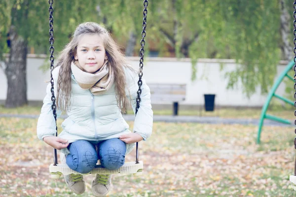 Cute Girl ubrana w dół kurtki siedzi na huśtawce na placu zabaw w chłodnym dniu jesiennym. — Zdjęcie stockowe