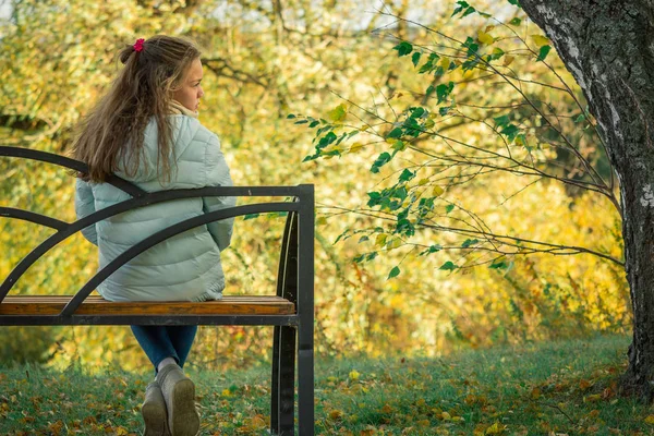 悲しい気分のライトダウンジャケットの美少女は、涼しい秋の日にベンチに座って休んでいます. — ストック写真