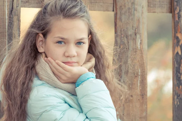 スカーフと青いダウンジャケットのかわいい女の子は、秋の涼しい日に村の木製のフェンスの背景に彼女の手を維持します. — ストック写真