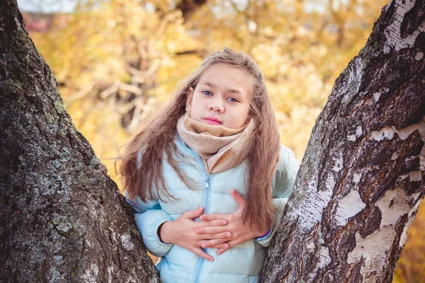 Glückliches Mädchen mit zerzausten Haaren in blauer Daunenjacke an einem Herbsttag an Birke gelehnt. — Stockfoto