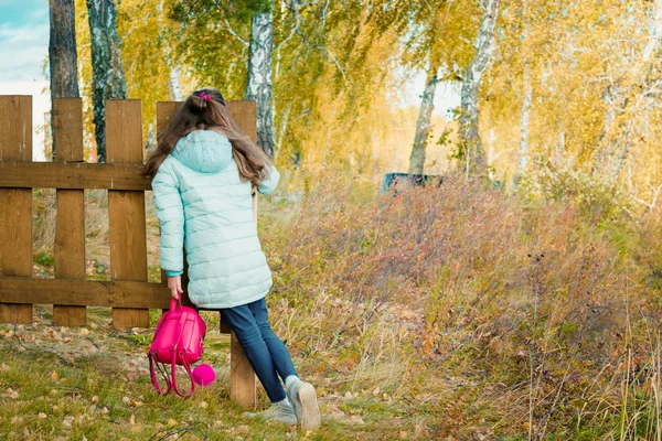 Ein großes Mädchen in Schal und blauer Daunenjacke hält an einem herbstlich kühlen Tag im Dorf einen roten Stadtrucksack am Holzzaun. — Stockfoto