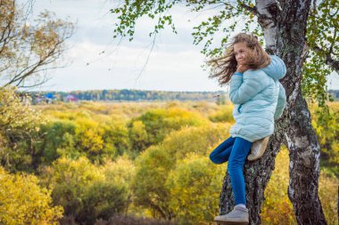 Contented girl with disheveled hair in blue down jacket sits on high birch tree on an autumn day. clipart