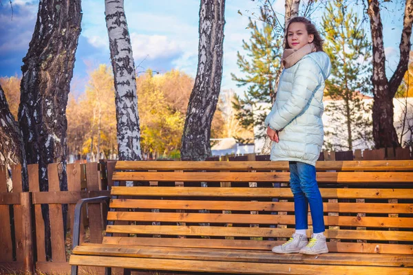 ライトダウンジャケットの美少女は涼しい秋の日にベンチに立つ. — ストック写真