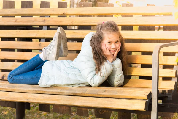 ライトダウンジャケットの魅力的な女の子は横たわってベンチに休み、涼しい秋の日に太陽を浴びて. — ストック写真