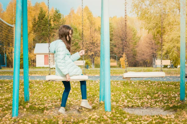 涼しい秋の日に遊び場でブランコに座ってダウンジャケットに身を包んだかわいい女の子. — ストック写真