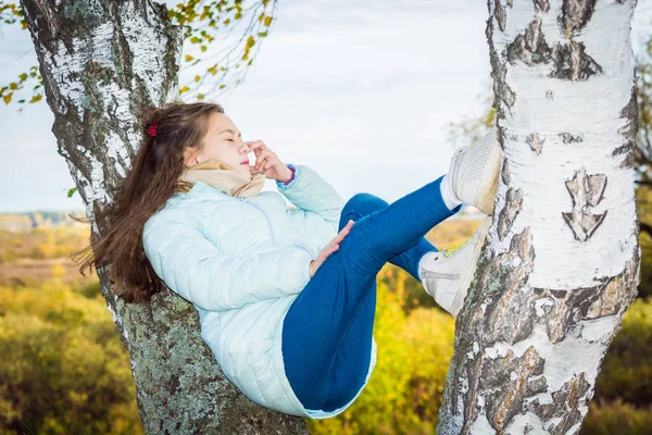 青いダウンジャケットの長い髪を持つ若い女の子は、秋の日に白樺の木に横たわっています. — ストック写真