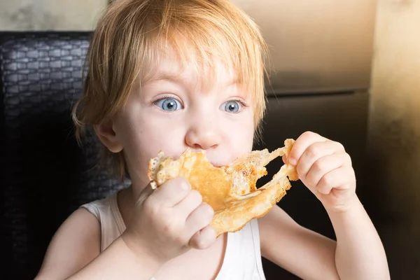 Bebê bonito com olhos azuis come deliciosas panquecas enquanto sentado na mesa da cozinha na cadeira. — Fotografia de Stock