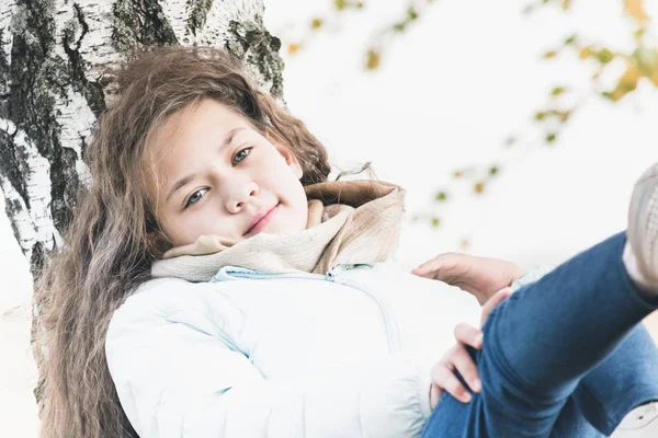 Молода дівчина з довгим волоссям у синій куртці лежить на березовому дереві в осінній день . — стокове фото