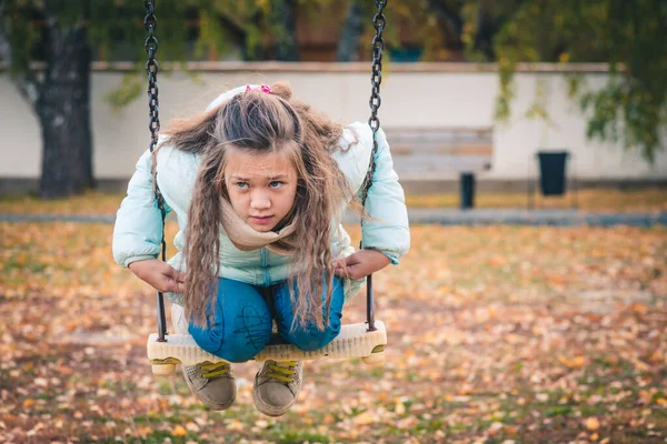 Nettes Mädchen in Daunenjacke sitzt an einem kühlen Herbsttag auf einer Schaukel auf dem Spielplatz. — Stockfoto