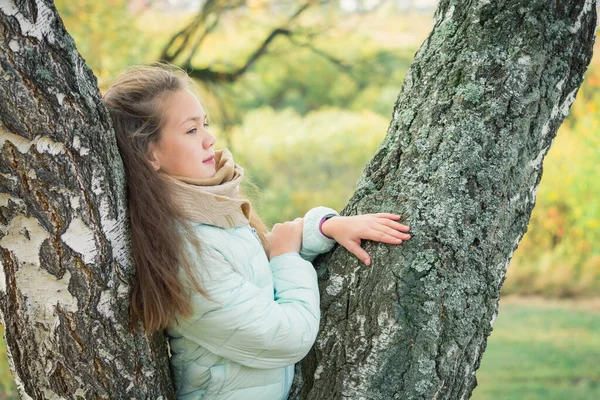 秋の日に白樺の木に寄りかかって青いダウンジャケットを着たペンシブな女の子. — ストック写真