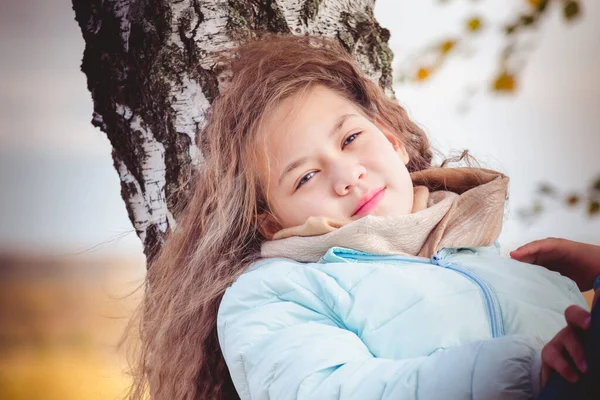 青いダウンジャケットの長い髪を持つ若い女の子は、秋の日に白樺の木に横たわっています. — ストック写真