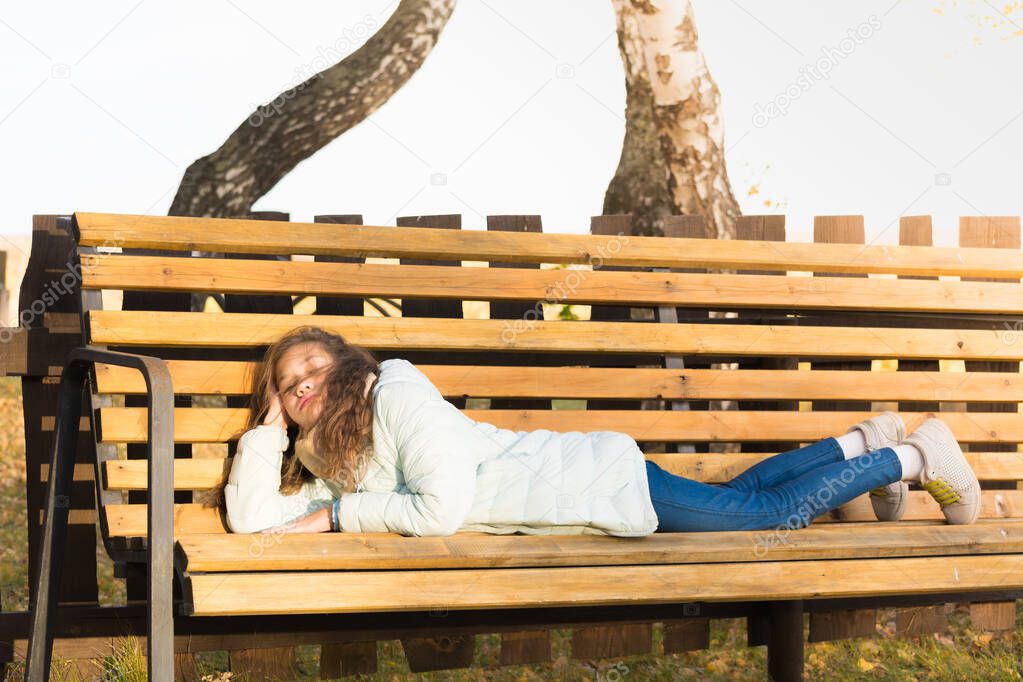 Charming girl in light down jacket lies and rests on bench, basking in the sun on cool autumn day.