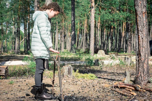 Girl with curls in light down jacket near burned out bonfire and stick for bowler hat in forest hike. — ストック写真
