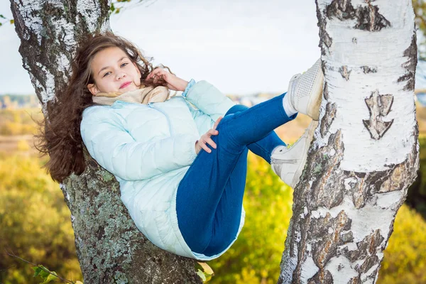 Junges Mädchen mit langen Haaren in blauer Daunenjacke liegt an einem Herbsttag auf Birke. — Stockfoto