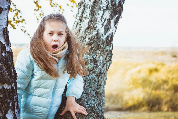 Girl with flowing hair in blue down jacket leaning against birch tree opened its mouth to the wind on an autumn day. — ストック写真